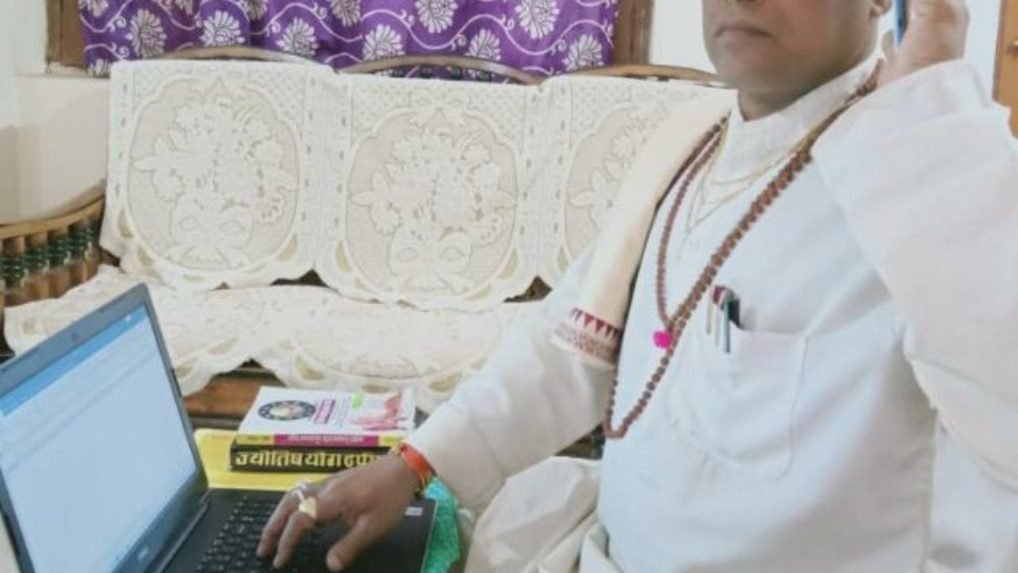 सफेद पोशाक पहने एक ज्योतिषी लैपटॉप और फोन के साथ बैठे नजर आ रहे हैं